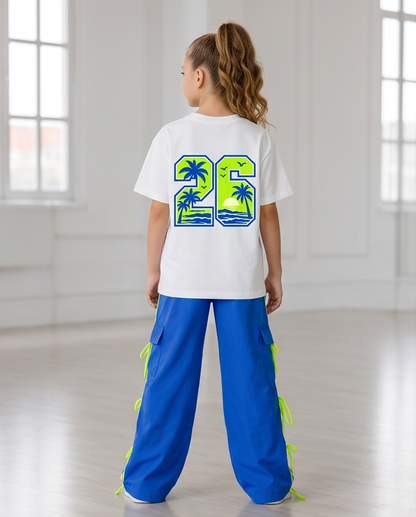 A child model viewed from behind wearing a white t-shirt with a large green and blue graphic on the back, paired with bright blue pants featuring neon green side ties, in a bright, sunlit studio.