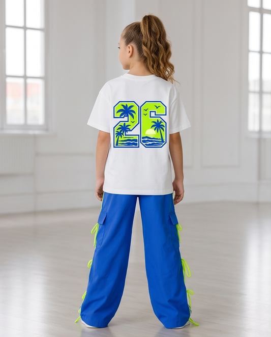 A child model viewed from behind wearing a white t-shirt with a large green and blue graphic on the back, paired with bright blue pants featuring neon green side ties, in a bright, sunlit studio.