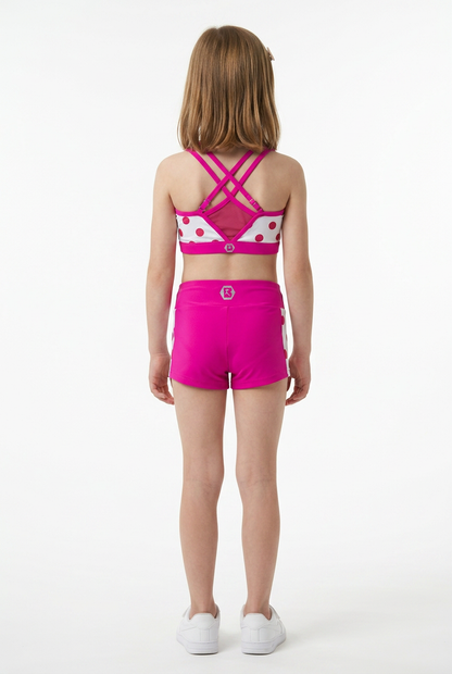 A young girl viewed from behind wearing a bright pink and white patterned sports bra and matching hot pink athletic shorts, standing against a plain white studio background.
