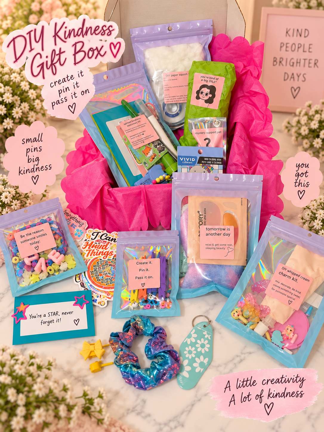 DIY Kindness Gift Box set arranged on a white marble surface with white flowers. The set includes a bright pink fabric basket, multiple zippered pouches with colorful contents, pastel pens, a scrunchie, a small keychain, and printed cards with kind-themed messages.