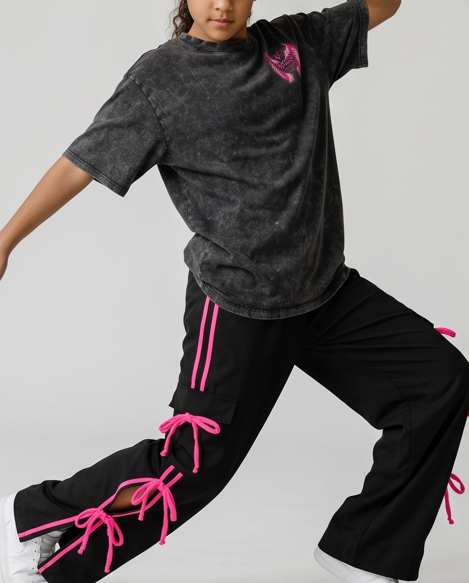 A child model wearing a washed black t-shirt with a pink graphic and black cargo pants featuring bright pink laces, posed dynamically against a light gray studio background.