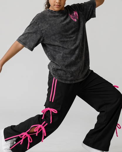 A child model wearing a washed black t-shirt with a pink graphic and black cargo pants featuring bright pink laces, posed dynamically against a light gray studio background.
